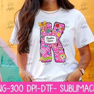 以下が含まれることがあります： 学校をテーマにしたグラフィックと「Maestra Karen」の文字が特徴の、大きなピンク色の文字「K」がデザインされた白いTシャツ。デザインには虹、鉛筆、リンゴが含まれています。画像の下部には「PNG-300 DPI-DTF- SUBLIMACIÓN」という文字があります。