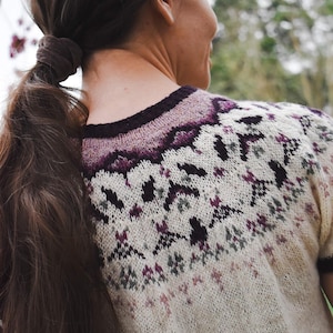 Può includere: Una donna indossa un maglione di lana bianco con un motivo geometrico viola e nero. Il maglione ha un collo alto e una vestibilità ampia.