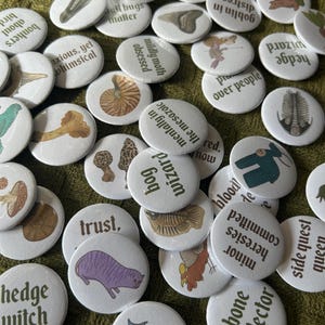 Könnte beinhalten: Eine Sammlung runder Ansteckbuttons mit verschiedenen Illustrationen, darunter Pilze, Fossilien, Tiere und Fabelwesen, jeweils kombiniert mit einzigartigen Textphrasen wie bog wizard, bone collector und side quest queen.