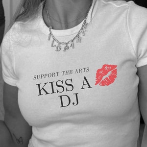 Puede incluir: Camiseta blanca con el texto "Support the Arts Kiss a DJ" y una impresi&oacute;n de labios rojos.