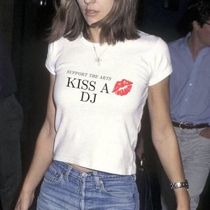 Puede incluir: Una camiseta blanca con el texto "Support the Arts Kiss A DJ" y una impresi&oacute;n de labios rojos.