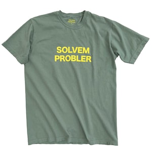 May include: A sage green t-shirt with the text "SOLVEM PROBLER" in yellow block letters across the front. The shirt has a crew neck and short sleeves. The text "SOLVEM PROBLER" is also printed on the back of the shirt.