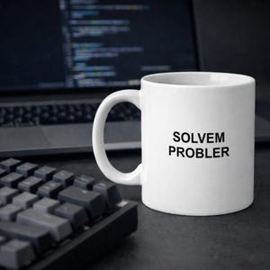 Op de afbeelding: Een witte keramische mok met de zwarte tekst "SOLVEM PROBLER". De mok staat op een donkere ondergrond naast een toetsenbord en een laptop.