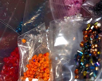 May include: A collection of small, colorful beads in clear plastic bags. The beads are various colors, including red, orange, yellow, blue, and iridescent. The bags are partially visible, creating a textured, layered effect.