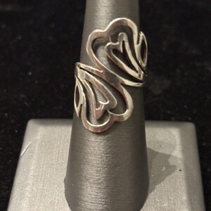 May include: A silver-colored ring with an openwork design featuring stylized heart and leaf shapes. The ring is displayed on a black ring holder. The ring's design is symmetrical and wraps around the finger.