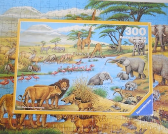 Könnte beinhalten: Ein 300-teiliges Ravensburger Puzzle mit einer afrikanischen Savannenlandschaft. Das Bild zeigt Giraffen, Elefanten, Löwen, Zebras und Flamingos an einem Wasserloch. Die Puzzledose ist gelb und hat das Ravensburger Logo.