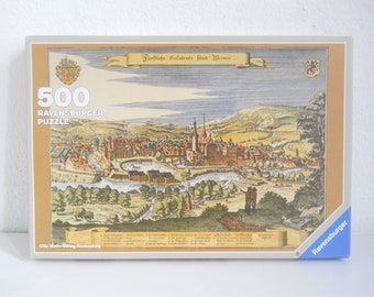 Könnte beinhalten: Eine 500-teilige Ravensburger Puzzle-Box mit einer antiken Illustration der Stadt Weimar, Deutschland. Die Schachtel ist beige mit einem blauen Ravensburger-Logo unten rechts. Das Bild zeigt eine Vogelperspektive der Stadt mit Gebäuden, Bäumen und einem Fluss.
