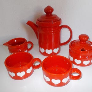 May include: A red ceramic tea set with white heart designs. The set includes a teapot, creamer, sugar bowl, and two cups. The teapot has a lid and a handle. The cups have handles. The sugar bowl has a lid with a small spoon opening.