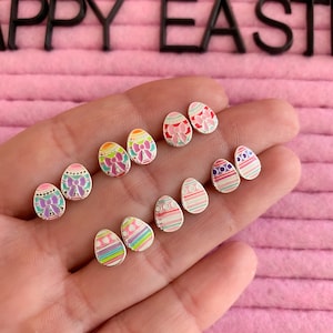 May include: A set of six pairs of silver earrings shaped like Easter eggs. The eggs are decorated with different colourful patterns, including stripes, polka dots, and bows.