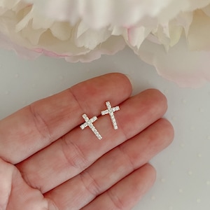May include: A pair of silver-colored cross stud earrings. Each cross is encrusted with small, clear crystals. The earrings are displayed on a light-colored surface, with a soft, blurred floral background.