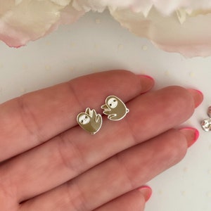 May include: A pair of silver-toned sloth stud earrings. Each earring features a brown sloth design with white and black details. The earrings are small and delicate, perfect for everyday wear.