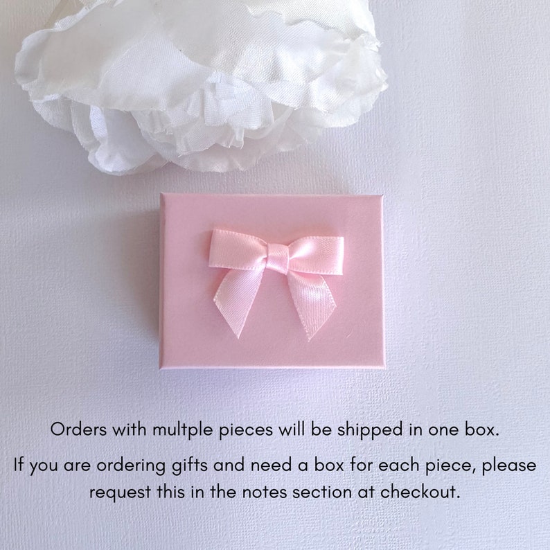 May include: A light pink gift box with a matching satin bow on top. A white, ruffled flower is in the upper left corner. The box is on a white background.