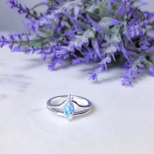 May include: A silver ring with a double band design. The ring features a pear-shaped opal and a small, round diamond. The ring is set against a backdrop of purple flowers.