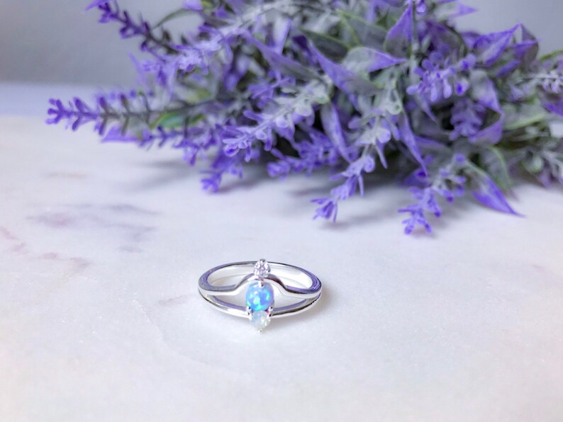 May include: A silver ring with a double band design. The ring features a pear-shaped opal and a small, round diamond. The ring is set against a backdrop of purple flowers.
