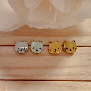 Puede incluir: Cuatro broches con forma de gato en tonos plateado, azul claro y dorado. Cada broche presenta un diseño minimalista de cara de gato con pequeños detalles. Los broches están dispuestos sobre una superficie de madera clara, con una tela blanca y arrugada en el fondo.