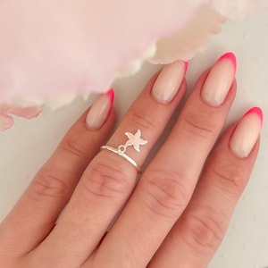 May include: A silver ring with a dangling starfish charm. The ring is worn on the middle finger of a hand with nude nails with pink tips.