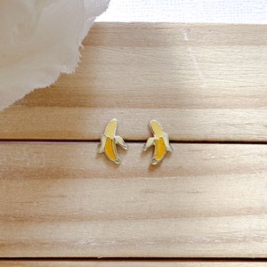 以下が含まれることがあります： バナナ型のスタッドピアス。各ピアスは、銀色の輪郭と緑色の先端が付いた黄色のバナナが特徴です。ピアスは明るい木製の表面に置かれています。