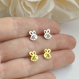 May include: Four small bee-shaped stud earrings. Two are silver-colored, and two are gold-colored. The earrings are displayed on a light background, showcasing their delicate design. The bee studs are a cute and simple accessory.