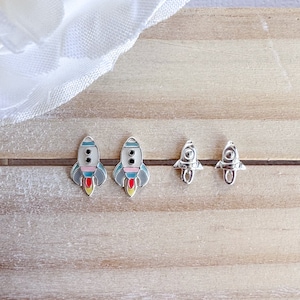 May include: A pair of silver rocket ship earrings with colorful details. The rockets are facing forward with a red flame coming out of the back. The earrings are on a wooden surface.