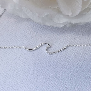 May include: A delicate silver necklace featuring a wave pendant. The pendant is a minimalist design, shaped like a stylized ocean wave. The chain is thin and connects to the wave at each end. The necklace is displayed on a textured white surface.