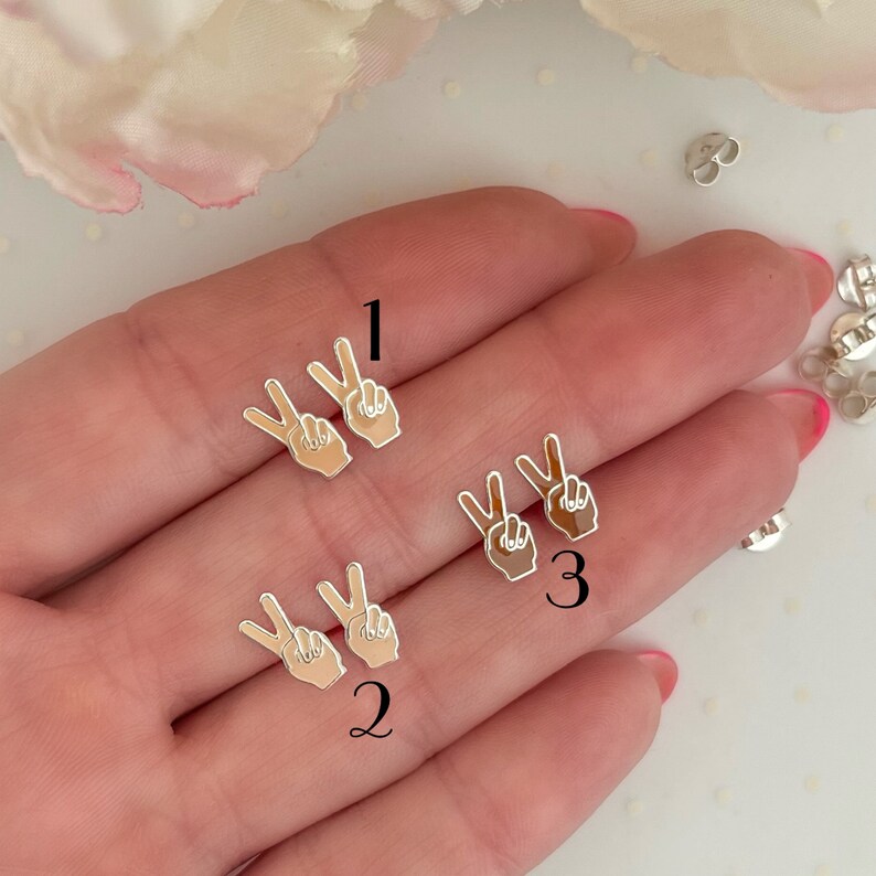 May include: Three pairs of peace sign earrings with different skin tones. The earrings are silver with a white enamel peace sign. The skin tones are light, medium, and dark.