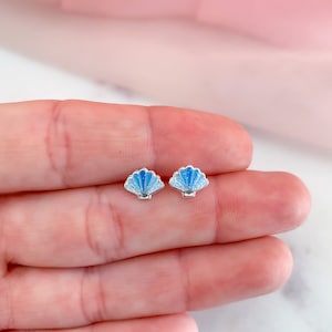 May include: A pair of small seashell stud earrings. Each earring features a blue and white enamel design on a silver-colored base. The earrings are shaped like scallop shells, with detailed ridges and a shimmering effect.
