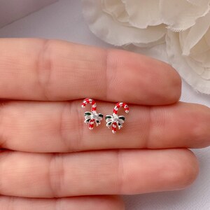 Puede incluir: Un par de pendientes de botón plateados con forma de bastón de caramelo. Cada pendiente presenta un diseño de bastón de caramelo a rayas rojas y blancas, un lazo verde y una pequeña gema transparente. Accesorio festivo.