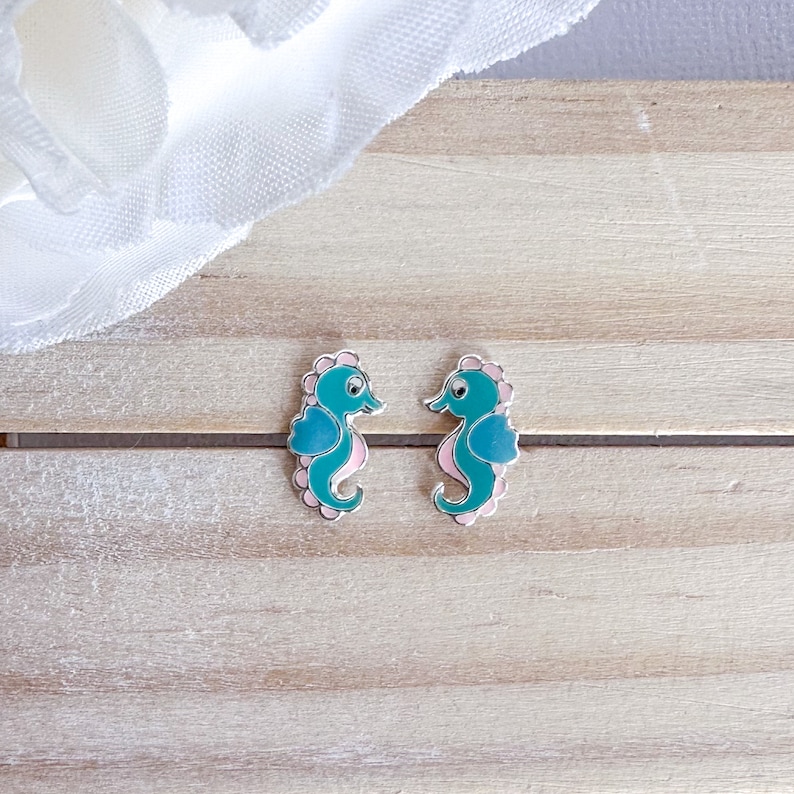 May include: A pair of silver stud earrings featuring a seahorse design. The seahorses are teal blue with pink accents.