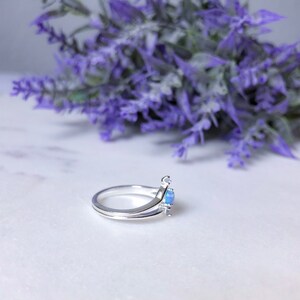 May include: A silver ring with a light blue gemstone. The ring has a unique design with a split band and a small accent on the side of the gemstone. The background features purple flowers.