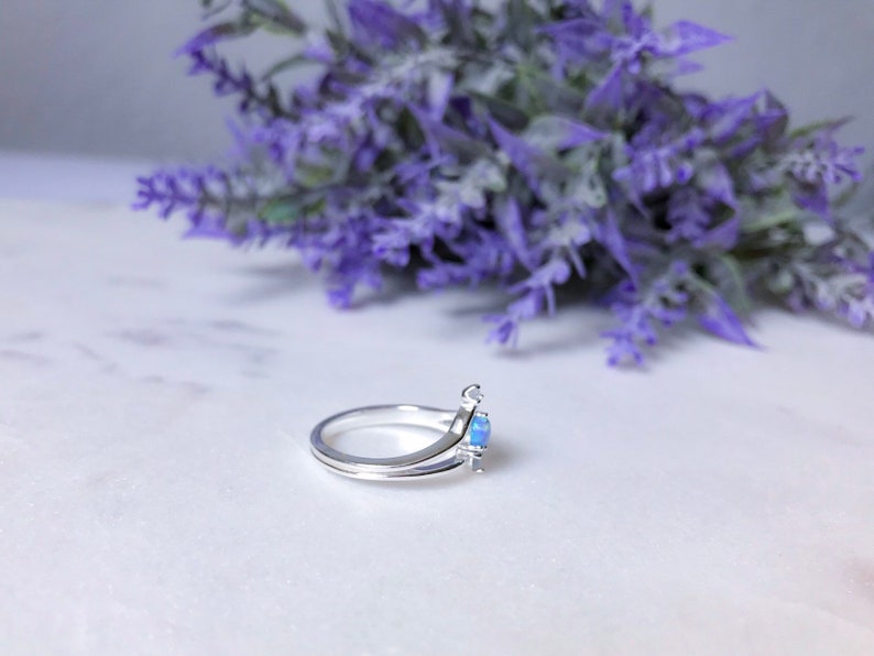 May include: A silver ring with a light blue gemstone. The ring has a unique design with a split band and a small accent on the side of the gemstone. The background features purple flowers.