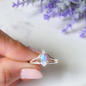 May include: A silver ring with a double band design, featuring a central oval-shaped opal stone and two smaller, clear accent stones. The opal has a blue and purple iridescent sheen. The ring is held against a soft background with lavender flowers.