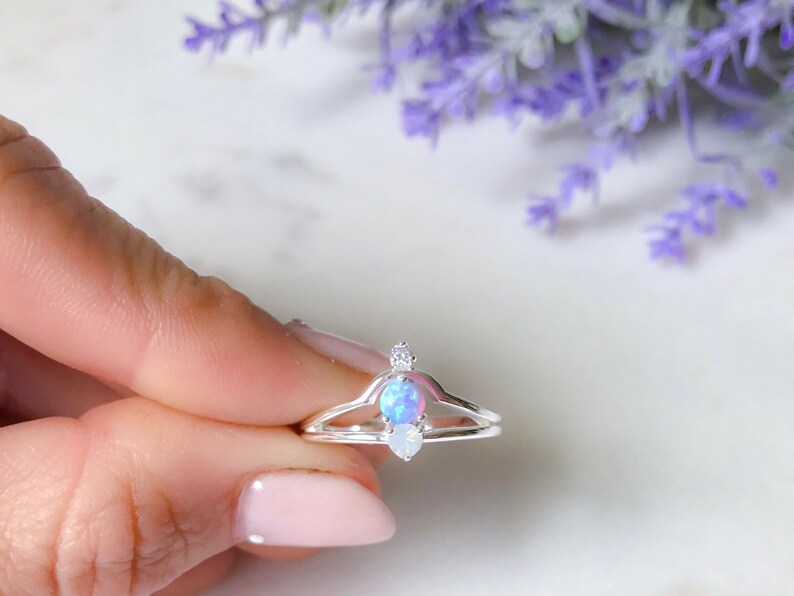 May include: A silver ring with a double band design, featuring a central oval-shaped opal stone and two smaller, clear accent stones. The opal has a blue and purple iridescent sheen. The ring is held against a soft background with lavender flowers.