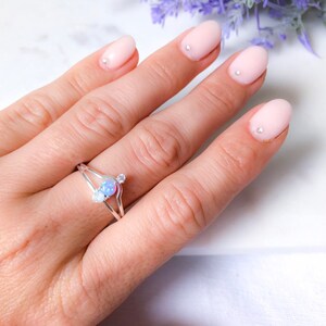 May include: A silver ring with a split band design, featuring a central, light blue opal-like stone and two smaller, clear stones. The ring is displayed on a hand with light pink nail polish and small silver nail accents. The background is blurred.