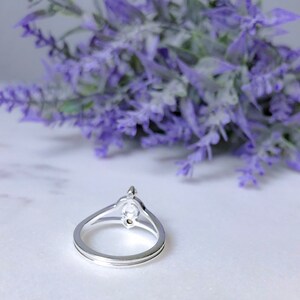 May include: A silver ring with a double band design. The ring features a central, oval-shaped clear stone, and a smaller stone above it. The ring is set against a backdrop of purple flowers.