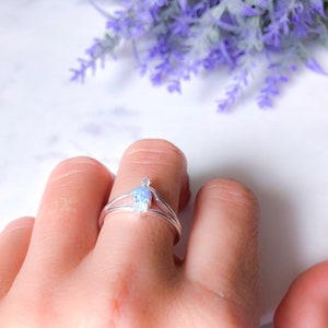 May include: A silver ring with a double band design, featuring a pear-shaped, iridescent gemstone. The ring is worn on a finger, with a backdrop of purple flowers.