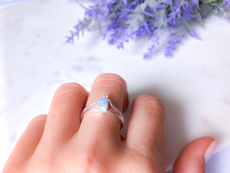 May include: A silver ring with a double band design, featuring a pear-shaped, iridescent gemstone. The ring is worn on a finger, with a backdrop of purple flowers.