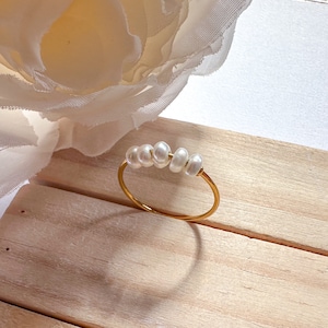 May include: A delicate gold ring featuring five irregular white pearls. The ring's band is thin and gold-colored, creating a minimalist and elegant design. The pearls are evenly spaced across the top of the ring.