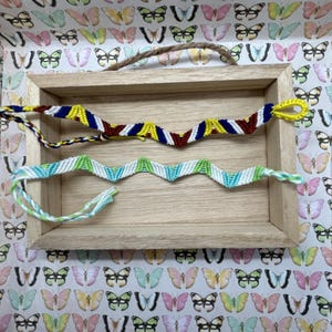 May include: Three friendship bracelets in a wooden frame. The bracelets are made with colorful threads and have different patterns. The top bracelet is yellow, red, blue, and white. The middle bracelet is green, white, and blue. The bottom bracelet is light green and white.