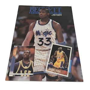 May include: A Beckett Basketball Monthly magazine featuring Shaquille O'Neal in an Orlando Magic jersey, with two basketball trading cards placed on top: one showing O'Neal in an LSU jersey and another in a Los Angeles Lakers uniform.