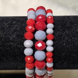 May include: A set of three stacked bracelets featuring faceted beads in alternating red and light gray colors. The beads are arranged in a repeating pattern, creating a textured appearance across the three individual strands of jewelry.