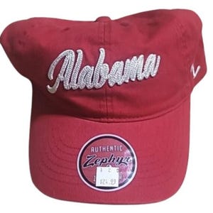 May include: A crimson baseball cap with the word "Alabama" embroidered in white script. The cap has a curved peak and a circular tag with the word "Authentic" and the brand "Zephyr".