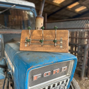 May include: A wooden coat rack with three metal hooks, each with a green ceramic knob. The rack is mounted on a weathered wooden board. The rack is resting on the bonnet of a blue Ford tractor.