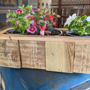 May include: A rectangular wooden planter box constructed from light-coloured, unfinished wood planks. The planter holds three dark plastic pots containing flowering plants: one with pink and yellow flowers, one with red fuchsia blooms, and one with white flowers.