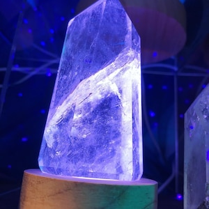 May include: A clear quartz crystal point on a wooden base with a light shining through it, creating a purple glow.