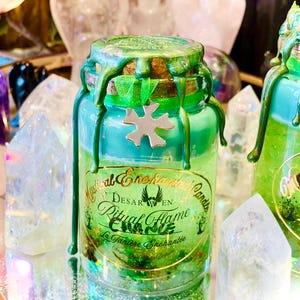 May include: A green glass jar candle with a green wax top and a gold label that reads "Mystical Enchanted Candles Desar en Ritual Flame Eva Marie La Tanniere Enchantée".