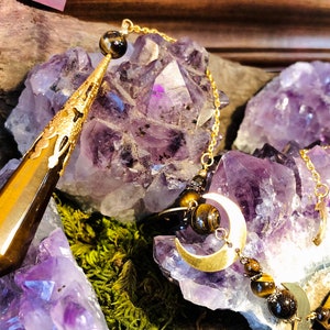 May include: A gold-toned pendulum with a brown and gold crystal point, hanging from a chain with a crescent moon charm and tiger's eye beads. The pendulum is resting on a cluster of amethyst crystals.