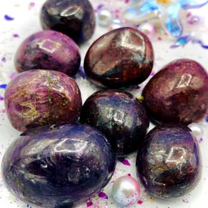 Può includere: Una collezione di pietre preziose lucide, di forma ovale, in tonalità viola intenso e bordeaux. Le pietre hanno una finitura lucida e motivi variabili, alcuni dei quali mostrano striature e inclusioni più chiare. La composizione include elementi decorativi come perle e glitter.