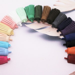 Op de afbeelding: Assortiment van 18 suède kwastjes in verschillende kleuren, waaronder roze, geel, blauw, groen, bruin, grijs, paars, oranje en rood. De kwastjes zijn in een halve cirkel op een wit oppervlak gerangschikt.