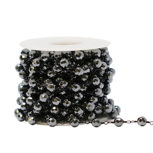 May include: A spool of black faceted beads on a white plastic spool. The beads are strung on a thin wire and are approximately 6mm in diameter. A short length of the beaded chain is extended from the spool.