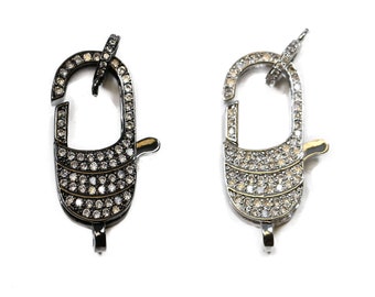 May include: Three jewelry clasps in different metallic finishes: black, silver, and gold. Each clasp is encrusted with small, clear gemstones. The clasps are oval-shaped with a hinged opening and a small loop at the bottom for attachment.